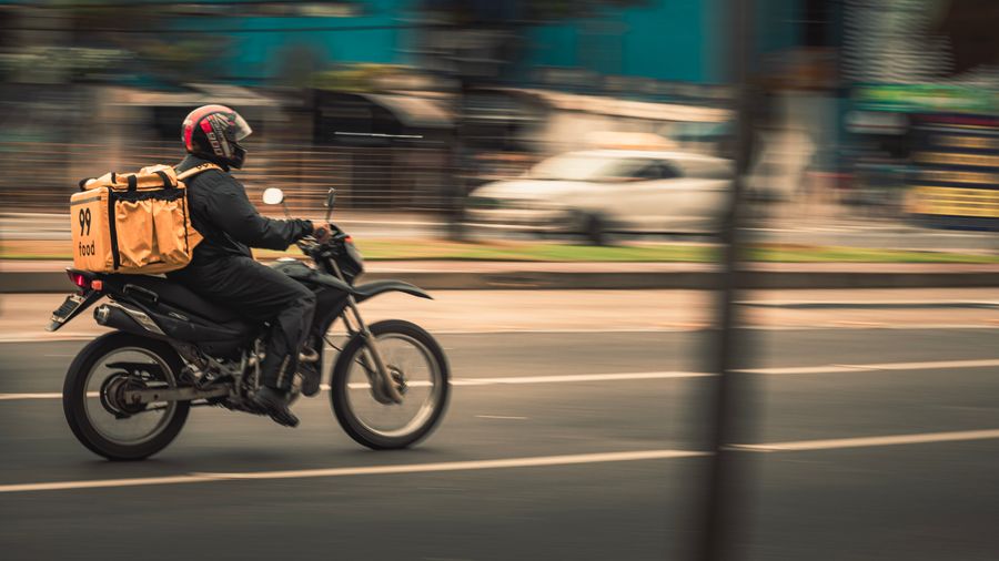 Motociclista com capacete e bag de entregas circulando por avenida urbana